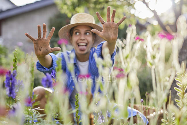 Fröhliche junge Frau zeigt schmutzige Hände, während sie inmitten von Pflanzen im Garten sitzt — Stockfoto
