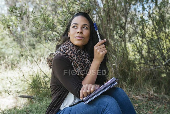 Nachdenkliche junge Frau schaut weg, während sie mit Tagebuch und Stift im Park sitzt — Stockfoto