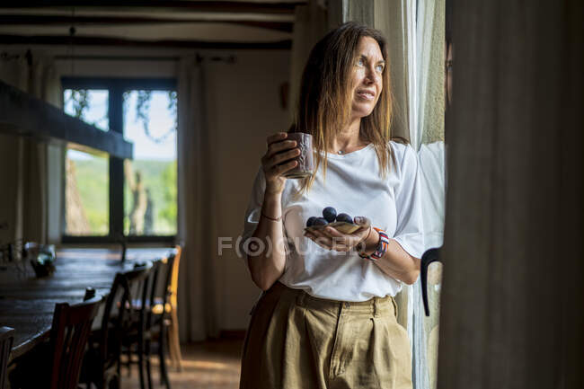 Задумчивая женщина-эксперт по еде держит в руках напитки и фрукты, глядя в окно — стоковое фото