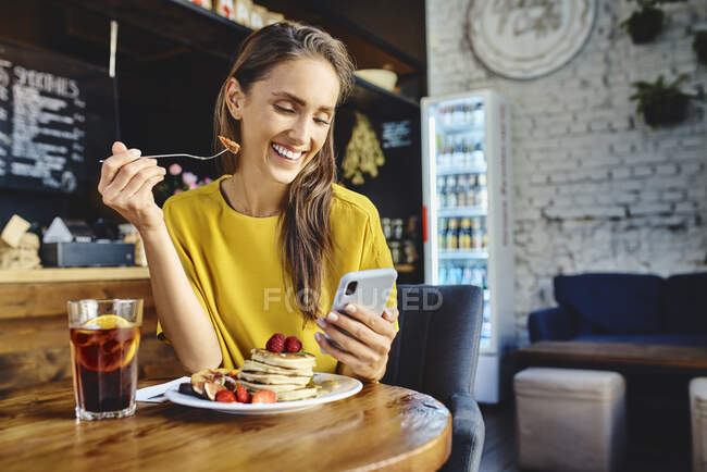 Smiling young female eating berry while using mobile phone at table in cafe — Stock Photo