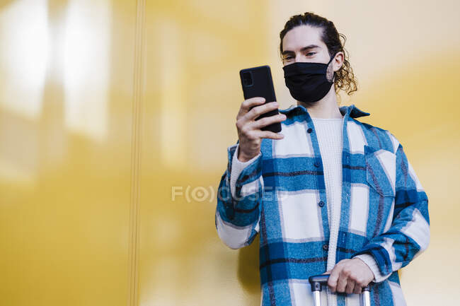 Giovane uomo che indossa maschera protettiva utilizzando il telefono cellulare mentre in piedi contro la parete gialla — Foto stock