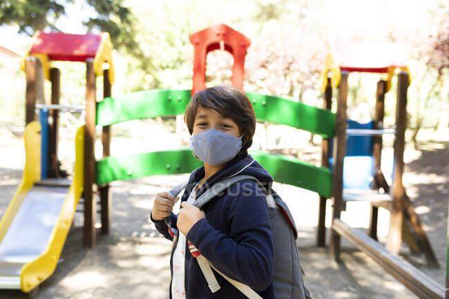 Boy wearing mask with backpack standing in schoolyard - foto de stock
