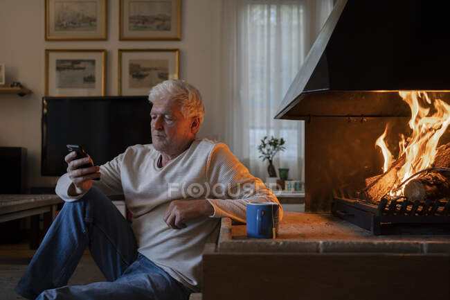 Hombre mayor usando el teléfono móvil mientras está sentado junto a la chimenea en casa - foto de stock