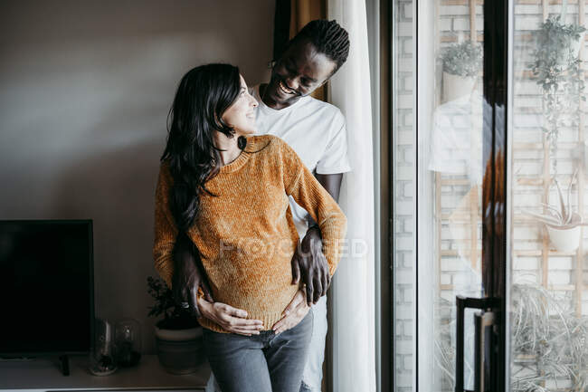 Happy young man looking at pregnant woman while standing by window at home — Stock Photo