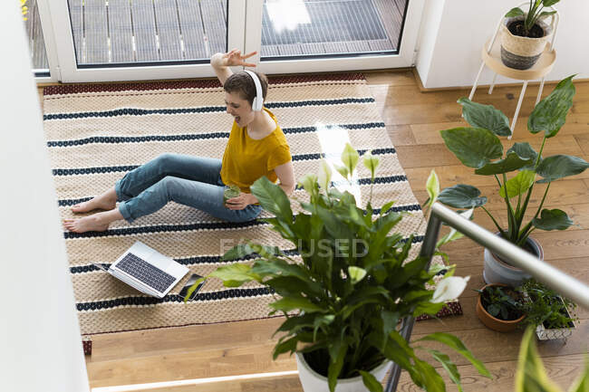 Cheerful woman listening music and dancing while sitting with laptop on carpet at home - foto de stock
