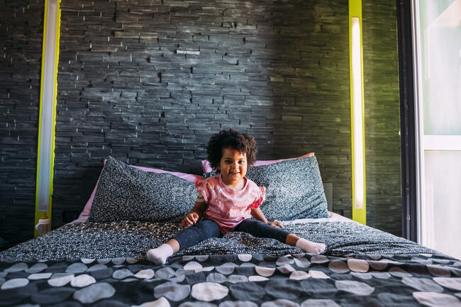 Bébé fille souriante avec jambes écartées assis sur le lit contre le mur à la maison — Photo de stock