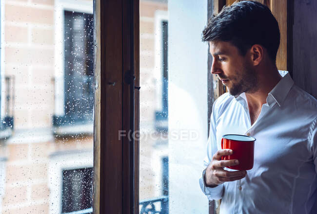 Jovem segurando copo olhando pela janela — Fotografia de Stock