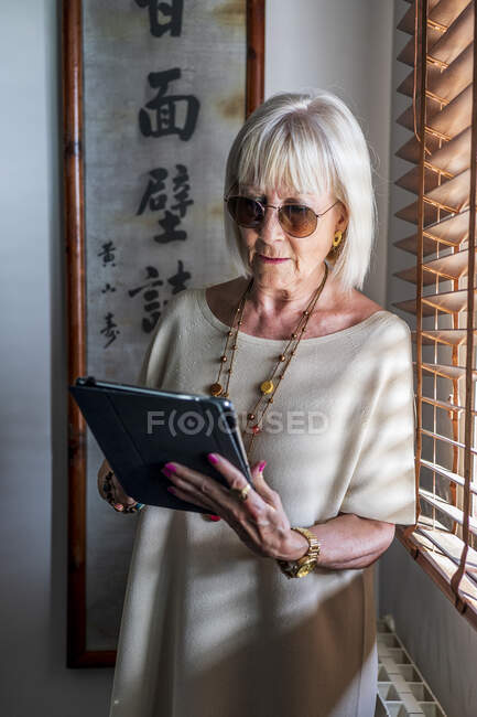Mujer mayor con gafas de sol usando tableta digital mientras está de pie junto a la ventana en casa - foto de stock