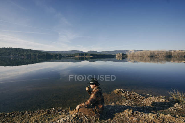 Bushcrafter beber taza de té en el lago en el bosque bajo el cielo azul - foto de stock