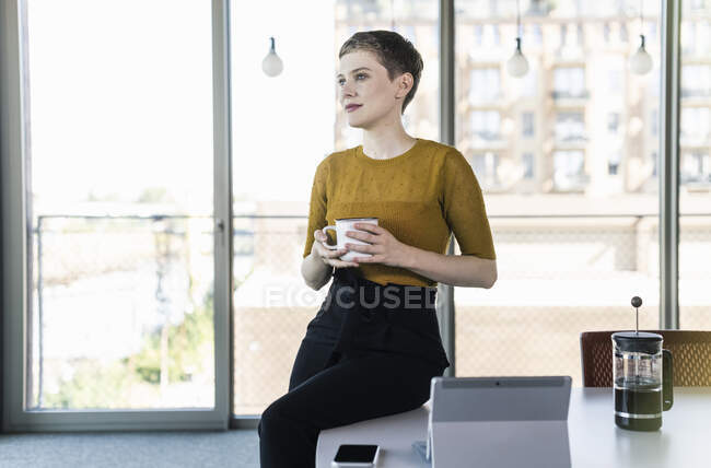 Деловая женщина сидит на столе в офисе держа кофейную кружку — стоковое фото