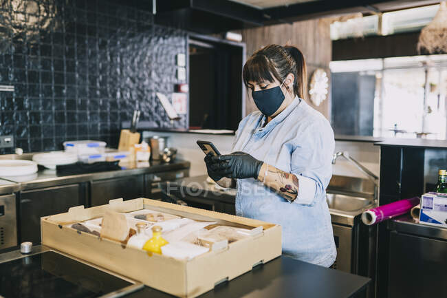Chef femminile utilizzando smart phone mentre in piedi da prendere fuori scatola di cibo al bancone della cucina nel ristorante durante coronavirus — Foto stock