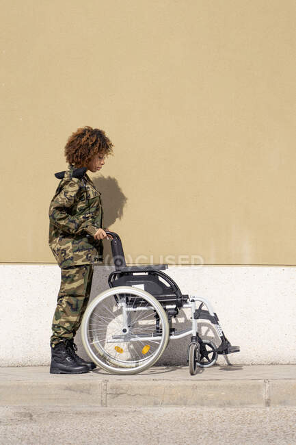 Mujer soldado del ejército mirando a la silla de ruedas vacía en el sendero contra la pared - foto de stock
