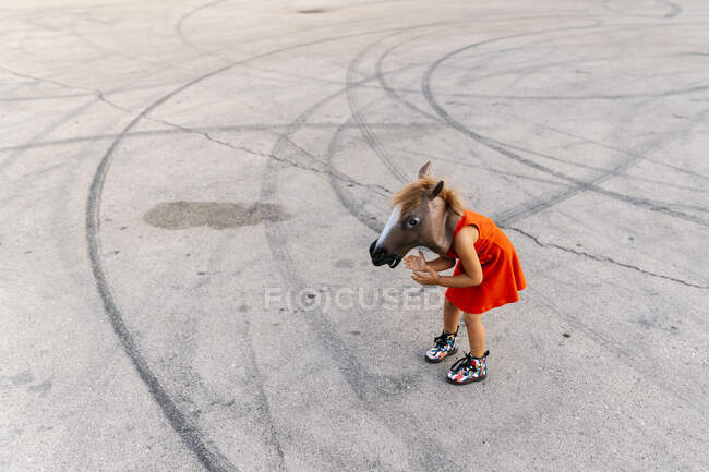 Bambina con testa di cavallo e vestito rosso, in piedi su asfalto con tracce di pneumatici — Foto stock
