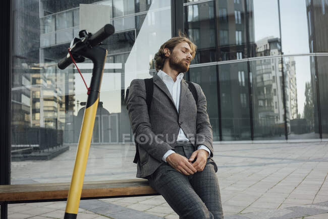 Tired male professional resting while napping on bench by electric push scooter in financial district — Fotografia de Stock