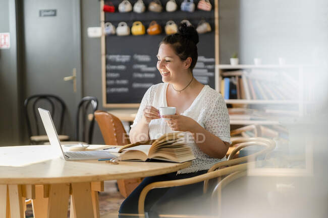 Sonriente mujer joven sosteniendo la taza de café mientras mira el ordenador portátil en la mesa en la cafetería - foto de stock