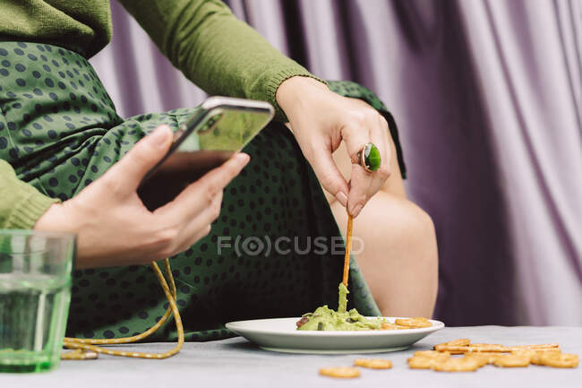 Primer plano de la mujer usando el teléfono inteligente mientras come guacamole y pretzel en la mesa en casa - foto de stock