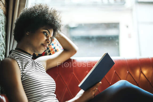 Femme adulte moyenne avec bouclé livre de lecture de cheveux tout en se relaxant sur le canapé dans le café — Photo de stock