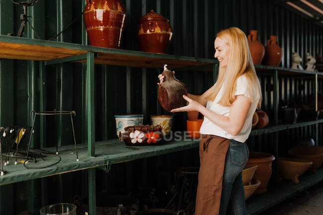 Mujer sonriente con cabello rubio sosteniendo cerámica mientras está de pie junto al estante en el taller - foto de stock