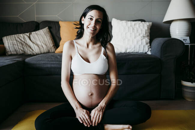 Mujer embarazada sonriente sentada en la esterilla de ejercicio en la sala de estar en casa - foto de stock