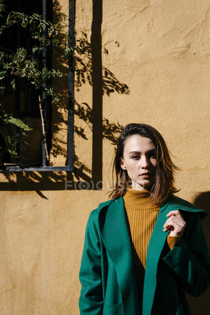 Schöne Frau in grüner Jacke steht an sonnigem Tag gegen Wand — Stockfoto