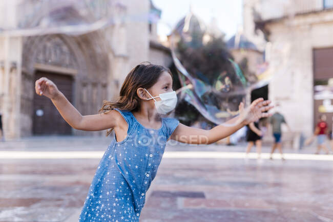 Carefree girl wearing mask running on street in city playing with soap bubble — Stock Photo