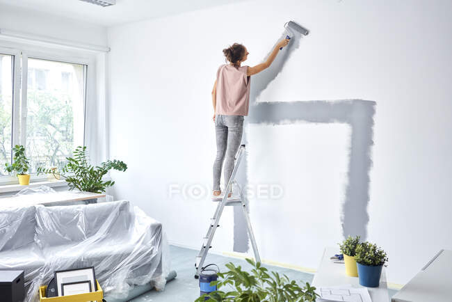 Giovane donna pittura parete con rullo del dolore mentre in piedi sulla scala a casa — Foto stock