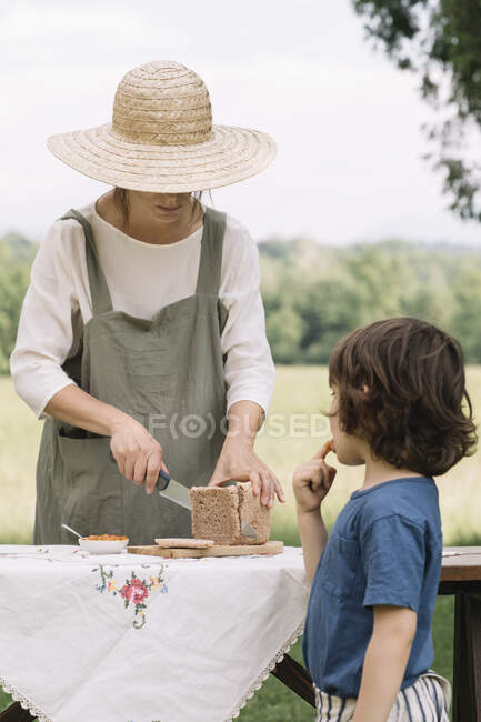 Donna taglio fetta di pane per ragazzo sul tavolo in giardino — Foto stock