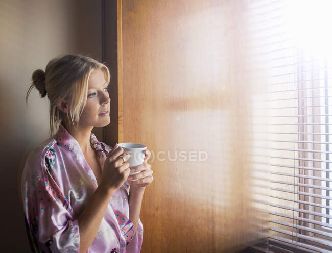 Jovem mulher pensativa em roupão de banho olhando através da janela enquanto toma café em casa — Fotografia de Stock