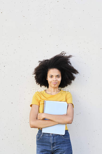 Souriant jeune étudiant tenant le livre tout en s'appuyant sur le mur — Photo de stock