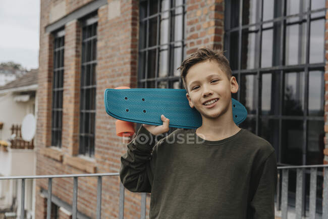 Ragazzo sorridente in posa con skateboard blu mentre in piedi contro la costruzione — Foto stock