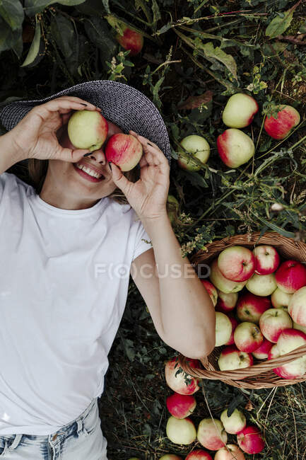 Femme souriante tenant des pommes devant les yeux alors qu'elle était allongée sur la terre ferme au verger — Photo de stock