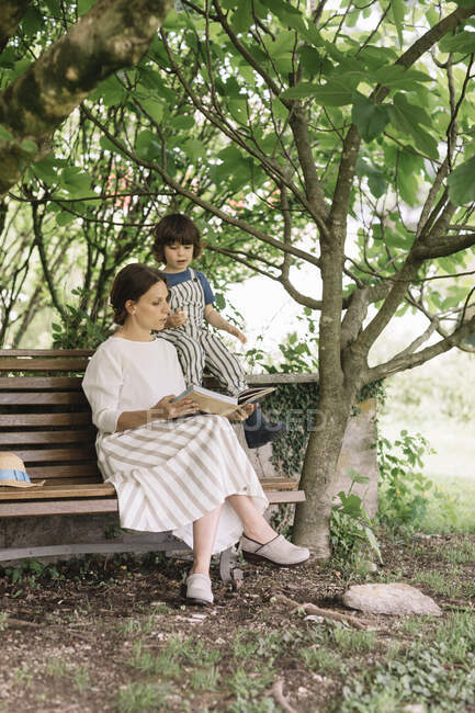 Beautiful woman and boy reading book while sitting on bench at backyard — Stock Photo
