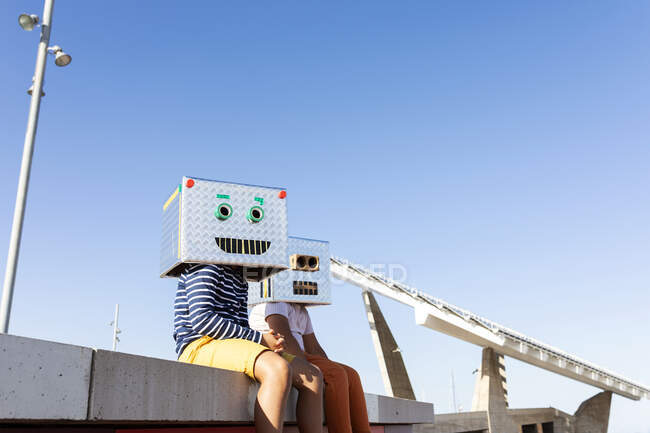 Boys wearing robot masks made of boxes while sitting on retaining wall against clear blue sky - foto de stock