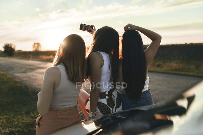 Amigas tomando selfie mientras están de pie en la carretera al atardecer - foto de stock