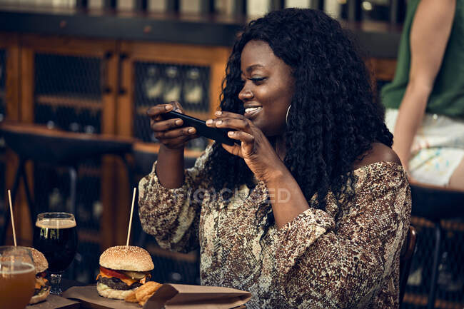 Mulher sorridente com smartphone tendo um hambúrguer em um pub — Fotografia de Stock
