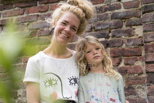 Sorridente donna bionda con figlia contro muro di mattoni nel cortile posteriore — Foto stock