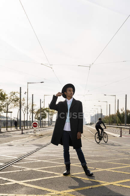 Giovane uomo guardando lontano mentre tiene il cappello tra binario ferroviario sulla strada — Foto stock