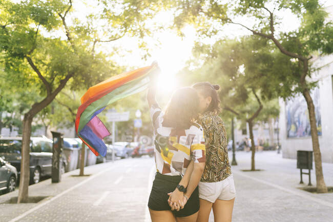 Giovane coppia lesbica baciare mentre sventola bandiera arcobaleno sul sentiero in città — Foto stock