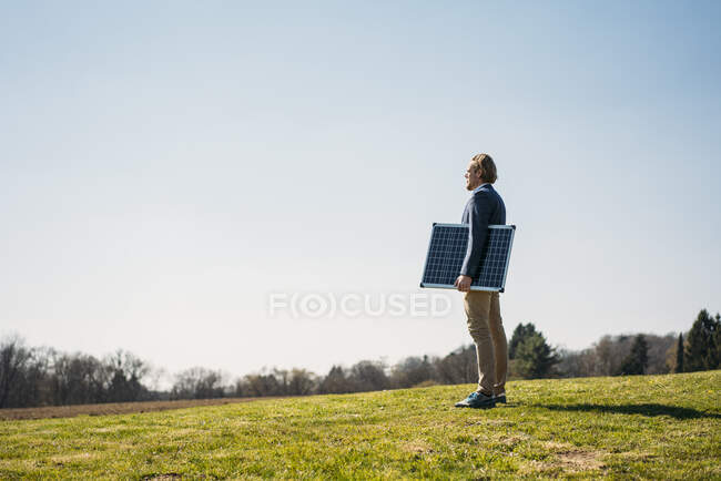 Empresario sosteniendo el panel solar mientras está de pie sobre la hierba y mirando hacia el parque contra el cielo despejado durante el día soleado - foto de stock