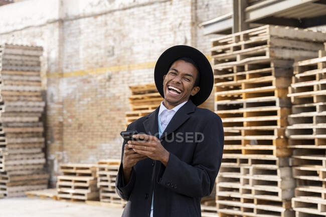 Junger Mann lacht, während er sein Handy gegen Holzpaletten hält — Stockfoto