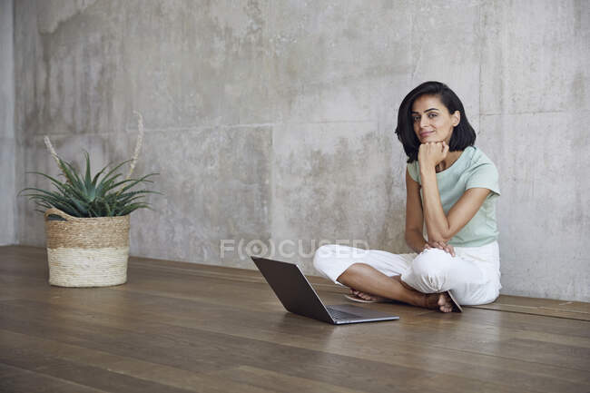 Imprenditrice sorridente seduta con laptop su pavimento in legno massello contro parete in ufficio — Foto stock