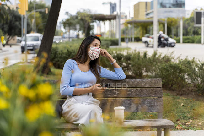 Betrachtende Frau, die während der COVID-19 auf einer Bank in der Stadt mit dem Smartphone wegschaut — Stockfoto