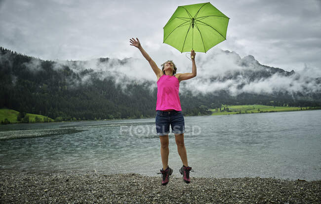 Felice donna matura che tiene l'ombrello mentre salta a Haldensee riva del lago durante la stagione delle piogge — Foto stock