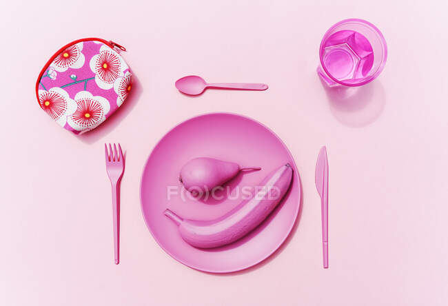 Estudio plano de vajilla de plástico rosa, vaso de agua, frutas rosadas y bolsita pequeña con patrón floral - foto de stock