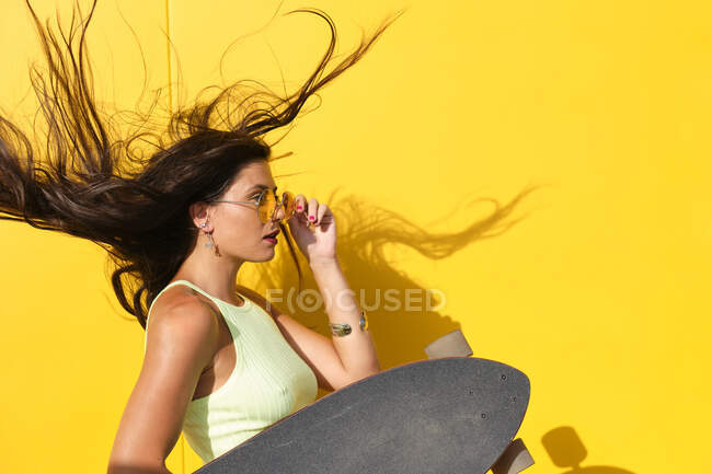 Portrait of young beautiful woman tossing hair in front of yellow wall with longboard in hand — Foto stock