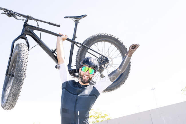 Athlète amputé masculin joyeux portant un vélo tout en se tenant debout contre un ciel clair — Photo de stock