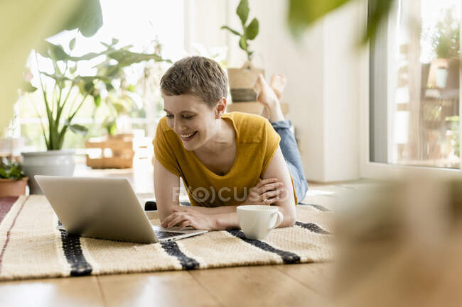 Smiling mid adult woman using laptop while lying on carpet at home — Foto stock