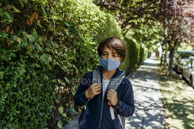 Schoolboy wearing mask looking away while standing by plants on footpath — Photo de stock