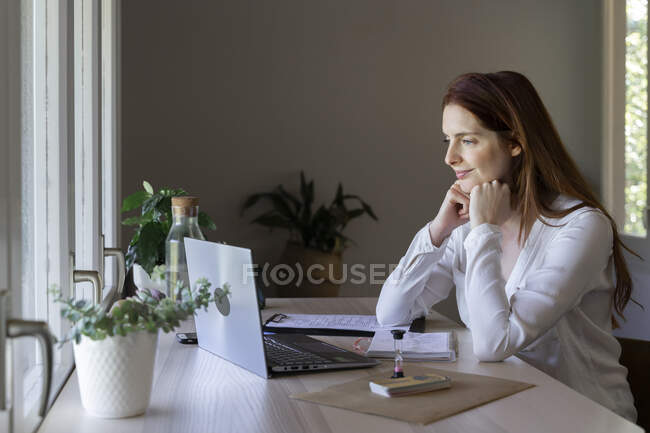 Medico femminile con mano sul mento che ascolta videochiamate sul computer portatile mentre è seduto a casa — Foto stock