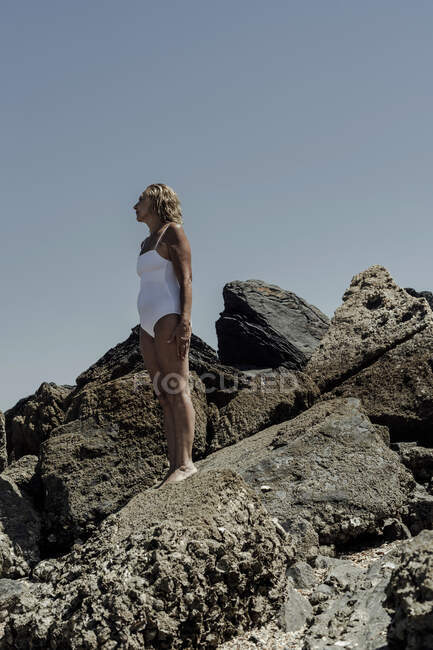Donna anziana che indossa costumi da bagno in piedi sulla roccia contro il cielo blu chiaro durante la giornata di sole — Foto stock